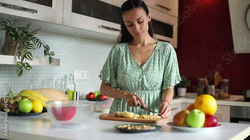 A woman in a green dress is slicing bananas on a wooden cutting board in a modern kitchen. Fresh fruit and a bowl are visible in the background. Healthy food preparation, cooking.