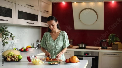 A woman in a green dress is chopping fruits on a kitchen counter, preparing a fruit salad. Fresh fruits, a bowl, and a modern kitchen setting are visible. Healthy eating, meal prep.