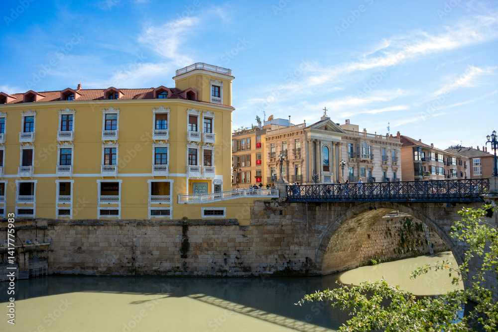 Obraz premium Puente de los Peligros over the Segura River, Murcia, Spain, with historic buildings in the background in bright afternoon light.