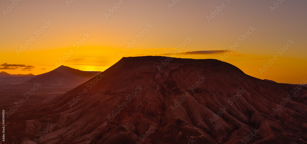 Fototapeta premium Beautiful aerial sunset image over Montana Roja volcanic crater silhouetted against the setting sun and skyscape near Corralejo, Fuerteventura, Canary Islands, Spain