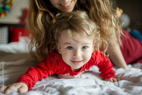 Joyful mother and curly-haired toddler wearing red shirts, playing together on a white bed, filled with happiness.