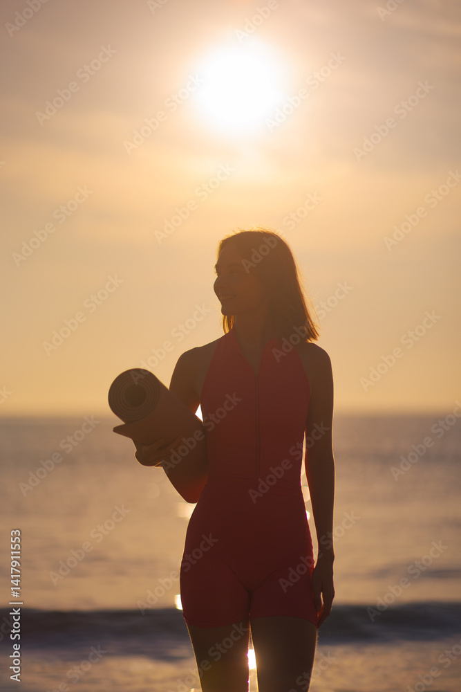 A silhouette of a woman in pink activewear holds a yoga mat while standing on the beach at sunset. The glowing sky and serene ocean backdrop create a peaceful and mindful atmosphere, perfect for