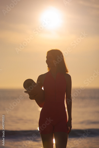 Obraz na płótnie A silhouette of a woman in pink activewear holds a yoga mat while standing on the beach at sunset. The glowing sky and serene ocean backdrop create a peaceful and mindful atmosphere, perfect for w salonie