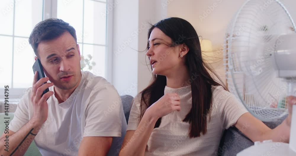 Family couple faces discomfort and trouble from hot summer weather at home living room, using fan for cool air in a challenging setting with focus on managing heat and relief every day.
