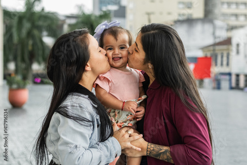 two Latina lesbian women, giving a kiss to their little daughter while holding her in their arms in the middle of the two, girl smiling very happy and joyful.