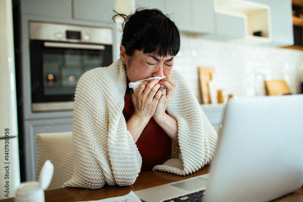 Sick woman blowing nose in tissue at kitchen table with laptop
