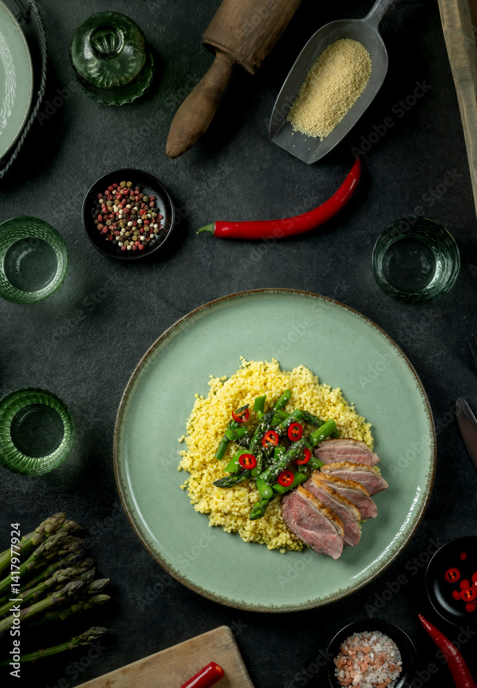 Roasted duck breast with asparagus, chili and couscous featuring kitchen utensils surrounding plate