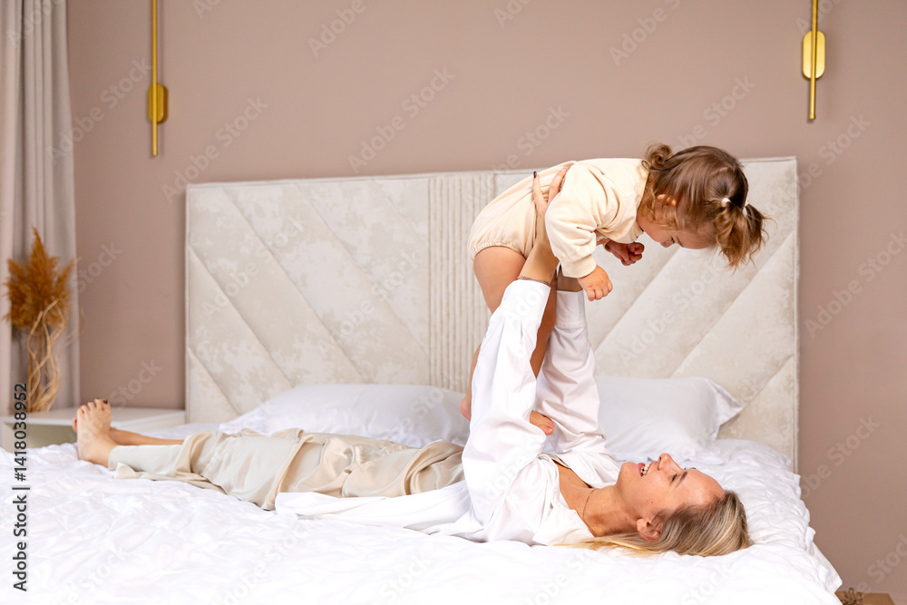 Dreamy flight. Carefree happy young mother play interact with cute beautiful baby in bright bedroom, lying on back on bed, holding laughing baby in air on outstretched arms. High quality photo