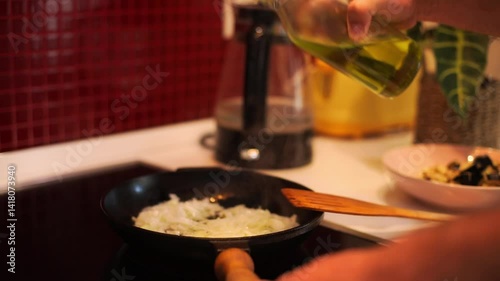 a person pouring olive oil into a frying pan containing sautéed onions. The scene is set in a modern kitchen with a warm, inviting atmosphere