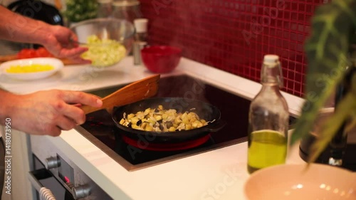 a person pouring chopped zucchini from a glass bowl into a frying pan with sautéed eggplant and onions. The cooking takes place on an induction cooktop in a modern kitchen
