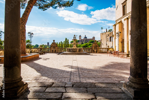 Large columns frame a view of Piazza Plebiscito in Viterbo, Italy, featuring a beautiful fountain, the Cathedral of San Lorenzo in the background, and red chairs adding a pop of color to the scene