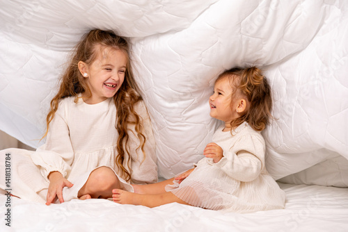 Obraz na płótnie Children sisters in bed under the blanket having fun, smiling. Happy little girl and her newborn baby sister hiding under the white blanket. Childhood concept. Copy space. High quality photo w salonie