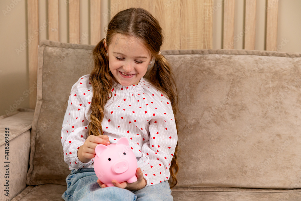Portrait of happy smiling little girl holding a pink piggy bank in hands while sitting on sofa in living room at home. Personal Savings. Child putting coin In piggybank. Copy space