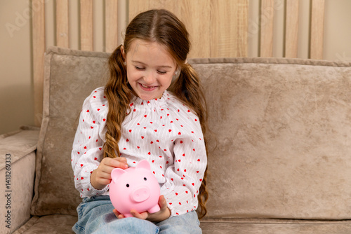 Obraz na płótnie Portrait of happy smiling little girl holding a pink piggy bank in hands while sitting on sofa in living room at home. Personal Savings. Child putting coin In piggybank. Copy space w salonie