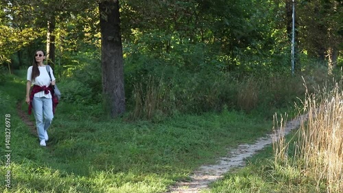 young woman walking on a scenic path in a park,wearing casual clothing with a backpack, enjoying sunny day outdoors. She smiles while strolling along the greenery, capturing the tranquility of nature