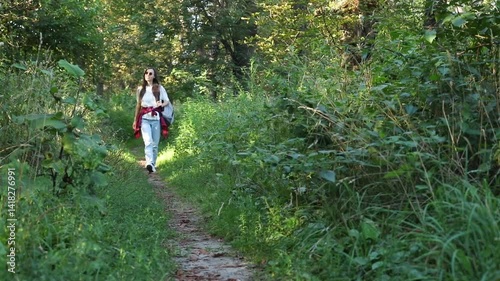 A young woman walks along a forest trail, dressed in casual jeans, a white T-shirt, and a plaid shirt tied around her waist. She carries a backpack, enjoying the nature walk in the sunlight.