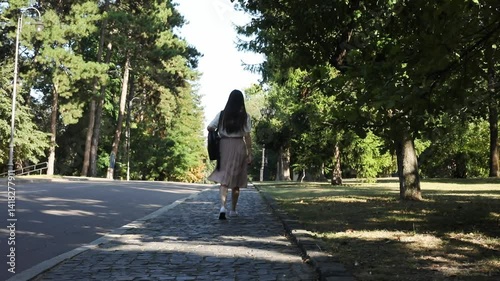 a woman walking along a peaceful park path on a sunny day. She wears a casual outfit with a light shirt, skirt, and a backpack, enjoying a leisurely stroll surrounded by greenery