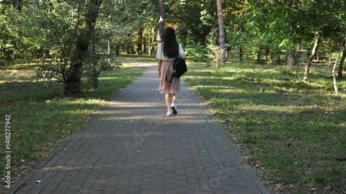 A young woman with a backpack walks along a paved path in a park surrounded by trees. She is casually dressed in a white top and pink skirt, enjoying a leisurely walk outdoors in nature.