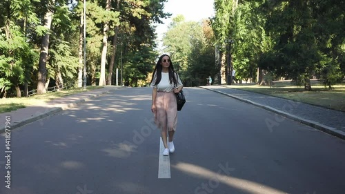 A young woman walking confidently along a tree-lined street, dressed casually in a white top and skirt, with sunglasses and a bag. The peaceful park setting adds a relaxed vibe to the scene.