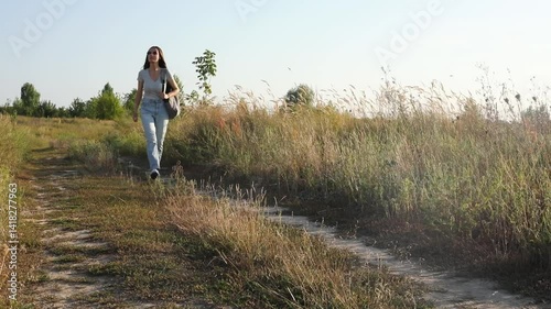 A woman walking on a dirt path in the countryside. She wears casual clothing, a backpack, and sunglasses, enjoying the peaceful nature surrounding her on a sunny day.