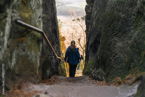 Φωτογραφία Solo hiker descending a narrow rocky passage with handrail in Sächsische Schweiz, framed by mossy sandstone cliffs and soft forest light
