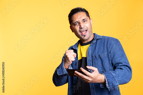 Pleased indian guy cheering after winning game on gaming console, standing against orange background. Delighted young adult bragging after being victorious in video gaming competition.