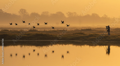 A photographer captures birds flying over a golden lake at sunrise, with reflections shimmering on the water and fog in the distance.