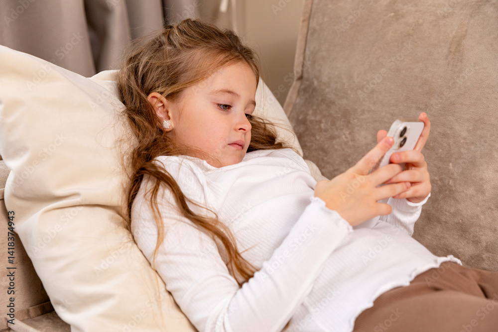 Cute little child lying on soft couch at living room, look at phone screen use game app contact mates at social network. Funny small girl engaged in watching online channel for children. Copy space