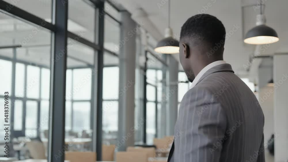 Back view of African-descent businessman walking through a modern office space, looking toward large windows, neutral tones. Back view of African-descent businessman walking through a modern office space, looking toward large windows, neutral tones.