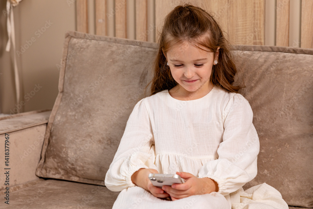 Smiling little child sitting on sofa at living room, look at phone screen use game app contact mates at social network. Funny small girl engaged in watching online channel for children. Copy space