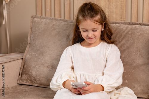 Obraz na płótnie Smiling little child sitting on sofa at living room, look at phone screen use game app contact mates at social network. Funny small girl engaged in watching online channel for children. Copy space w salonie