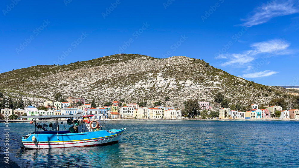 Fototapeta premium Colorful Waterfront Houses And Blue Fishing Boat In A Calm Bay, Peaceful Coastal Town At The Foot Of A Rocky Hill, Bright Sunny Day, Seaside Tranquility, Mediterranean Island Life