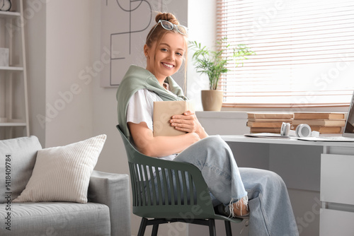 Smiling young woman holding book at home desk, remote learning and education,...