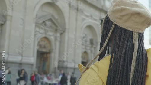 Black Latina woman with braids, yellow jacket, beret, and sunglasses, standing amazed as the camera circles 360° around her, sightseeing in a big city with a mix of modern and classical architecture