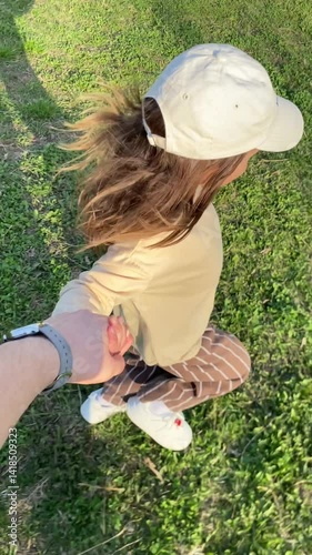 vertical video First-person view of holding hands with a little girl child wearing a white baseball cap, long-sleeved shirt, striped trousers, and white sneakers, walking on a grassy field on a sunny 