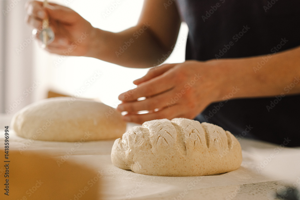 Obraz premium Woman hands skillfully score the surface of a round dough loaf, preparing sourdough bread for baking. The bright kitchen setting provides a warm atmosphere and fresh ingredients.