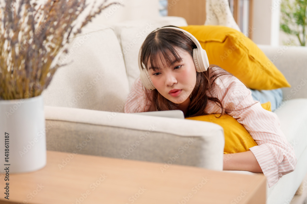 young woman is relaxing on couch while wearing headphones and watching something on her laptop. She appears focused and comfortable, surrounded by soft pillows in cozy living space.freeelancer young woman is relaxing on couch while wearing headphones and watching something on her laptop. She appears focused and comfortable, surrounded by soft pillows in cozy living space.freeelancer