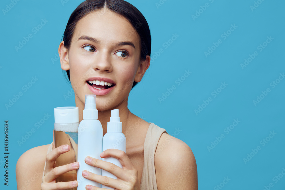 Young woman with a joyful expression holding skincare products against a blue background, promoting healthy skincare routines and beauty concepts Young woman with a joyful expression holding skincare products against a blue background, promoting healthy skincare routines and beauty concepts