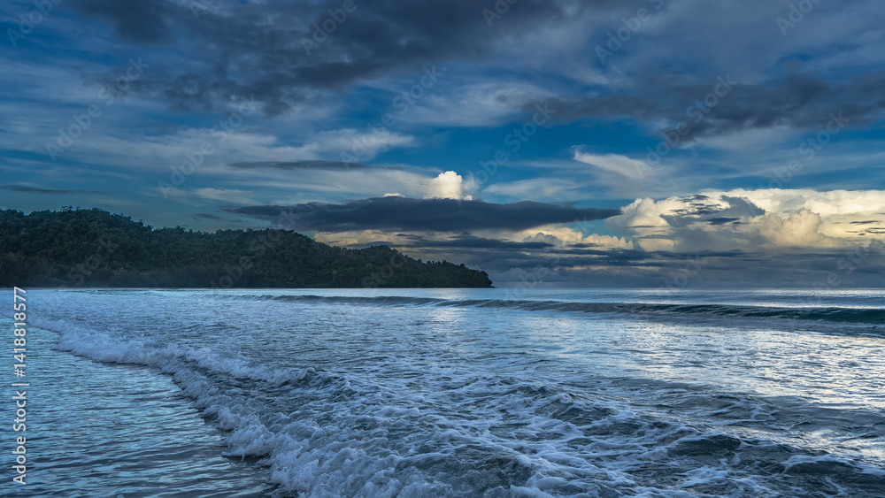 Fototapeta premium Morning by the ocean. The waves roll towards the shore, foam, and spread out. The clouds in the blue sky are highlighted in gold. A hill in the distance. Malaysia. Borneo. Kota Kinabalu.