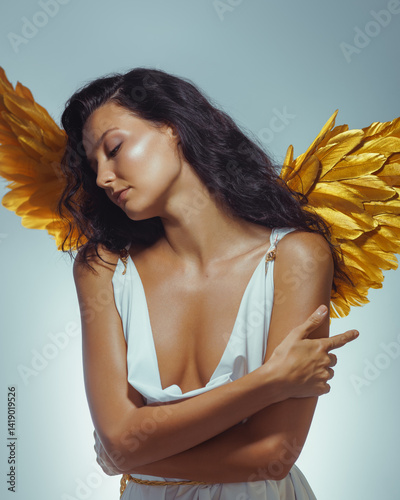 Obraz na płótnie Beautiful young Caucasian model with golden angel wings gently embracing herself in a white Greek-style dress. Conceptual and emotional fashion photo ideal for beauty, fantasy, or editorial use. w salonie