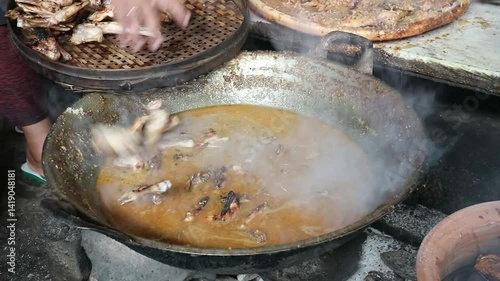 The process of cooking chicken by simmering it with various spices in a large pan over a wood stove in a simple and traditional kitchen.

