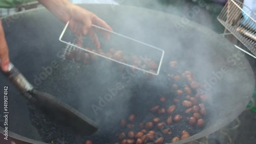 A market vendor roasts chestnuts in quartz sand with brown sugar, releasing aromatic smoke into the air, sifts through a stainless steel basket to separate the chestnuts from the sand. photo