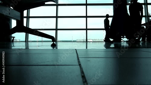 Passengers wait to board in the airport departure hall. Their silhouettes are visible against large windows in the waiting area. Slow motion video. Passengers wait to board in the airport waiting area