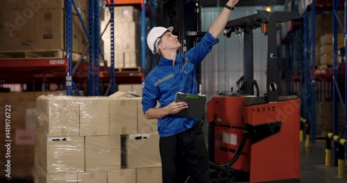 Caucasian male warehouse supervisor holds clipboard and points upward during forklift check while reviewing storage area logistics and documenting observations in cargo environment with industrial