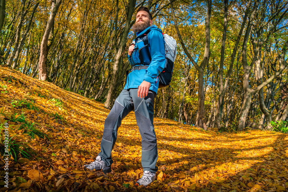 Low-angle view of a bearded male hiker in a blue jacket and backpack walking through an autumn forest blanketed in golden leaves. Sunlight creates long shadows on the ground. Low-angle view of a bearded male hiker in a blue jacket and backpack walking through an autumn forest blanketed in golden leaves. Sunlight creates long shadows on the ground.