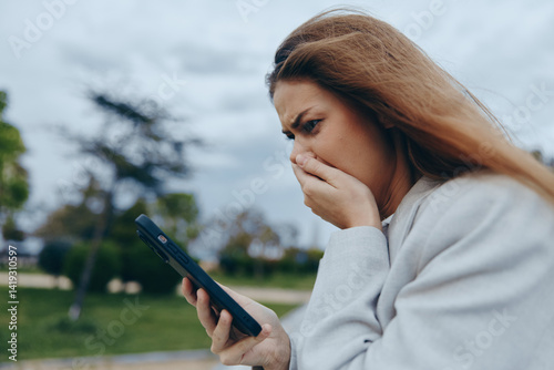 Obraz na płótnie Worried woman looking at phone screen outdoors with anxious expression, emotional stress, casual clothing, cloudy sky background, financial crisis concept w salonie