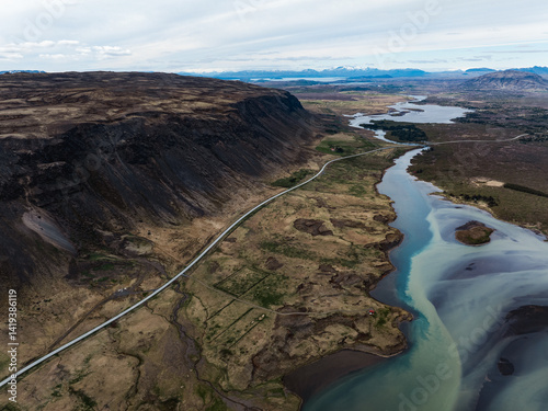 Aerial view of a winding river and road skirting a steep dark cliff, with glacial waters and a distant mountain range under a cloudy Icelandic sky.