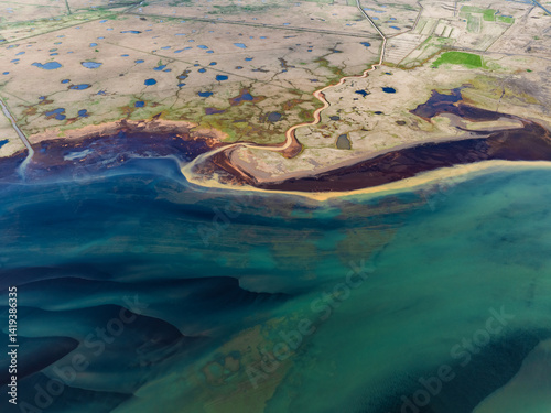 Aerial shot of a coastal plain filled with blue ponds, a winding river, and colorful sediments flowing into the turquoise sea, forming a natural abstract masterpiece.