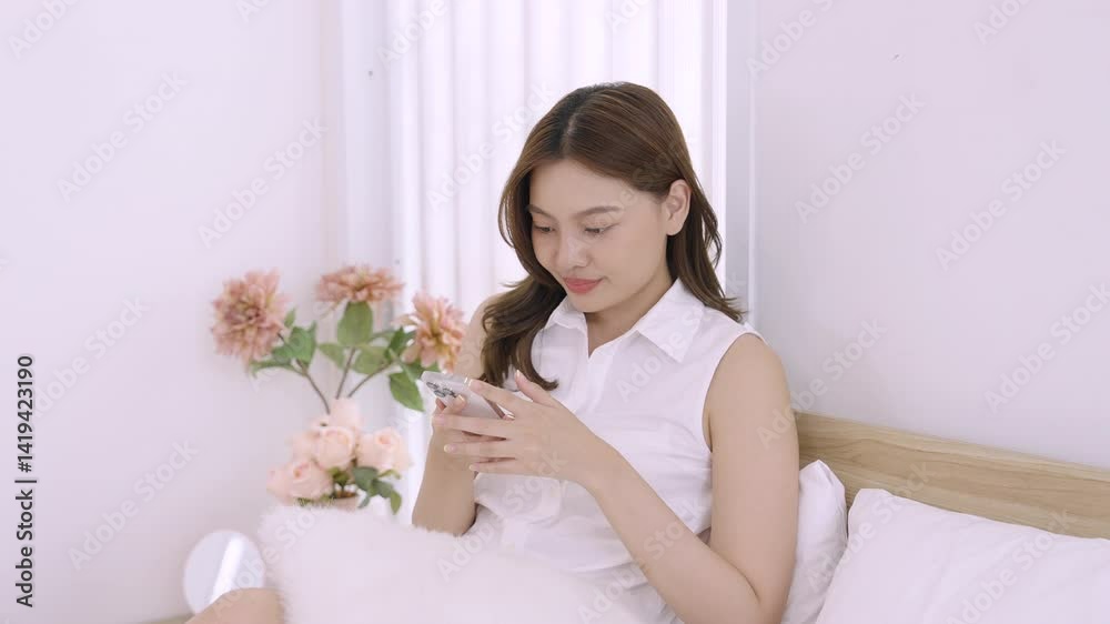 Asian young woman sitting on bed holding smartphone with soft smile looking down during peaceful morning moment surrounded by floral decoration in cozy bedroom expressing calm joy Asian young woman sitting on bed holding smartphone with soft smile looking down during peaceful morning moment surrounded by floral decoration in cozy bedroom expressing calm joy