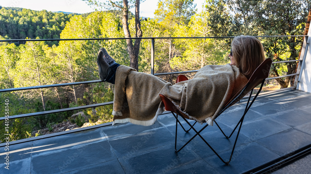 Woman lying on a chair with a blanket wrapped up and enjoying the views of the forest from the terrace.